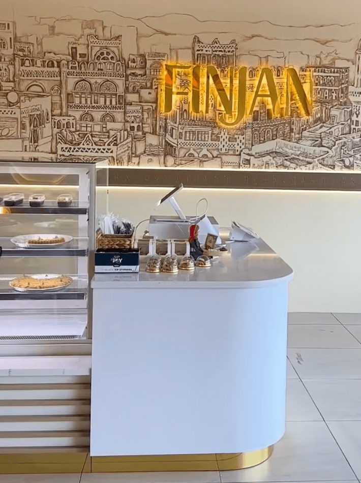 Finjan Cafe interior: counter, pastry case, and gold Finjan sign on a sketched mural wall